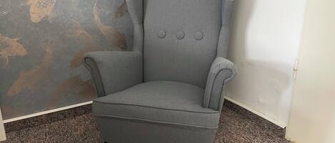 A grey armchair with a high back and armrests, positioned in a corner against a wall with a fish pattern.
