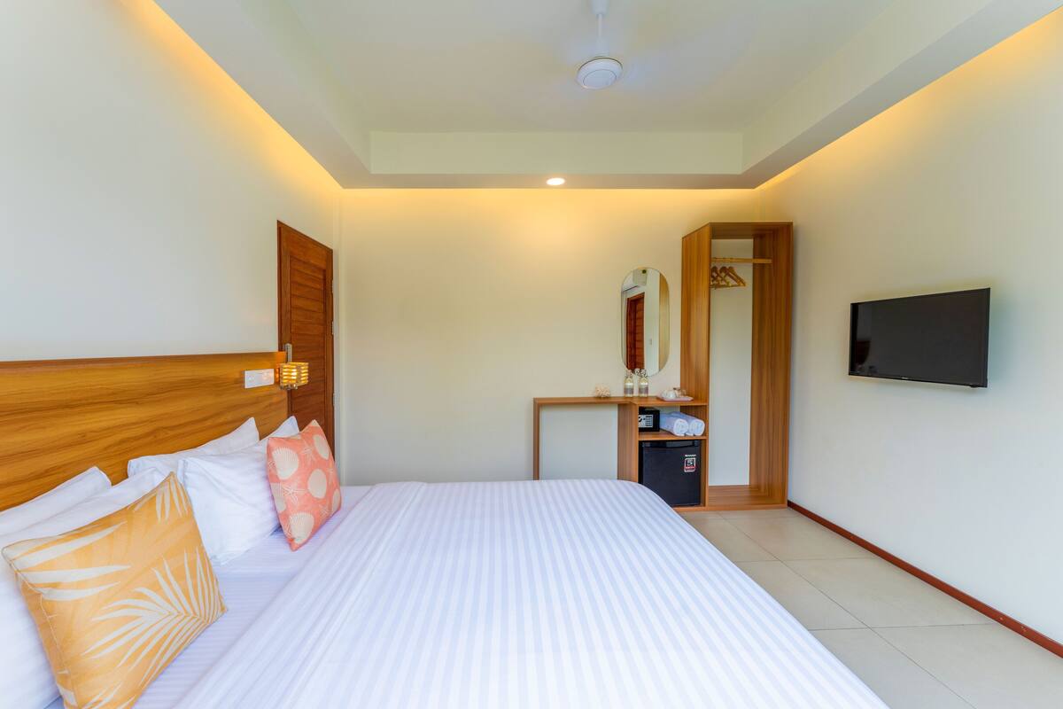 A hotel room with a large bed, a wooden headboard, a bedside table with a mirror, a TV mounted on the wall, and a small refrigerator.