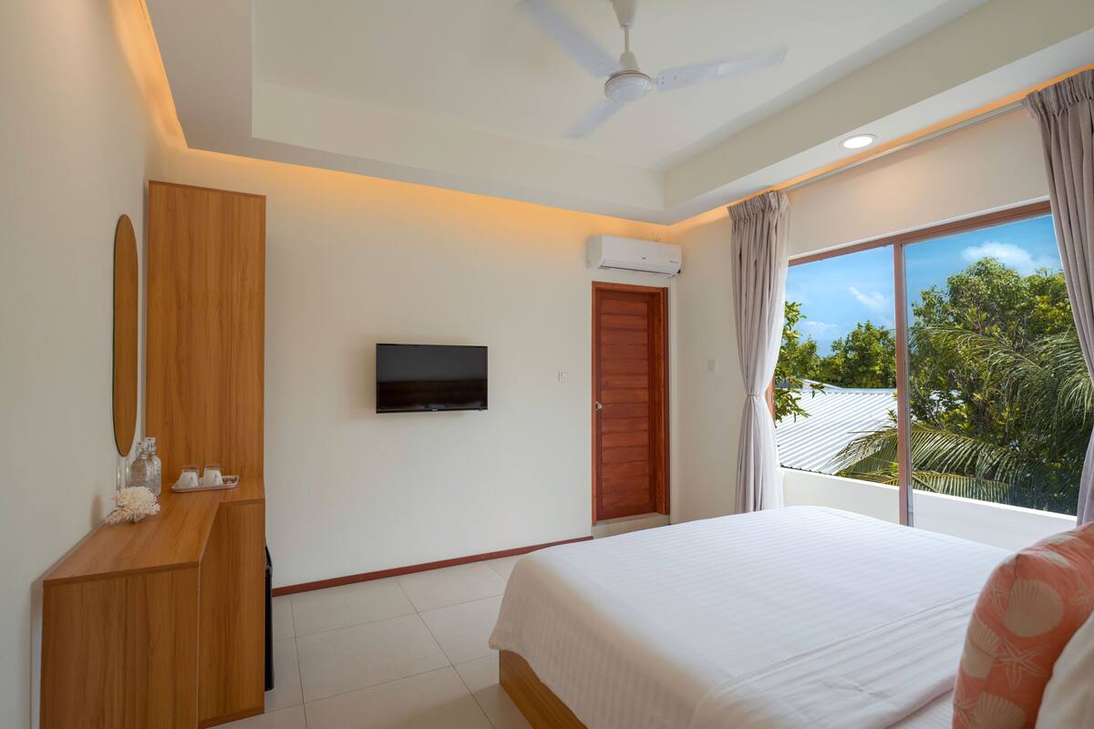 A bedroom with a bed, a TV, a wooden dresser, and a window with a view of trees.