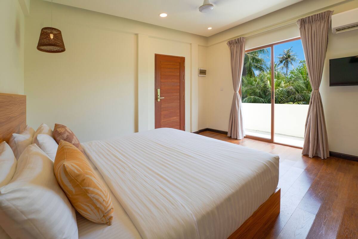 A bedroom with a large bed, a wooden headboard, a ceiling fan, and a view of palm trees through the window.