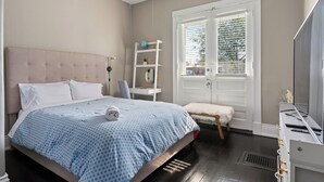 Ein Schlafzimmer mit Bett, Schminktisch, Stuhl, Hocker und Fenster mit Jalousien.