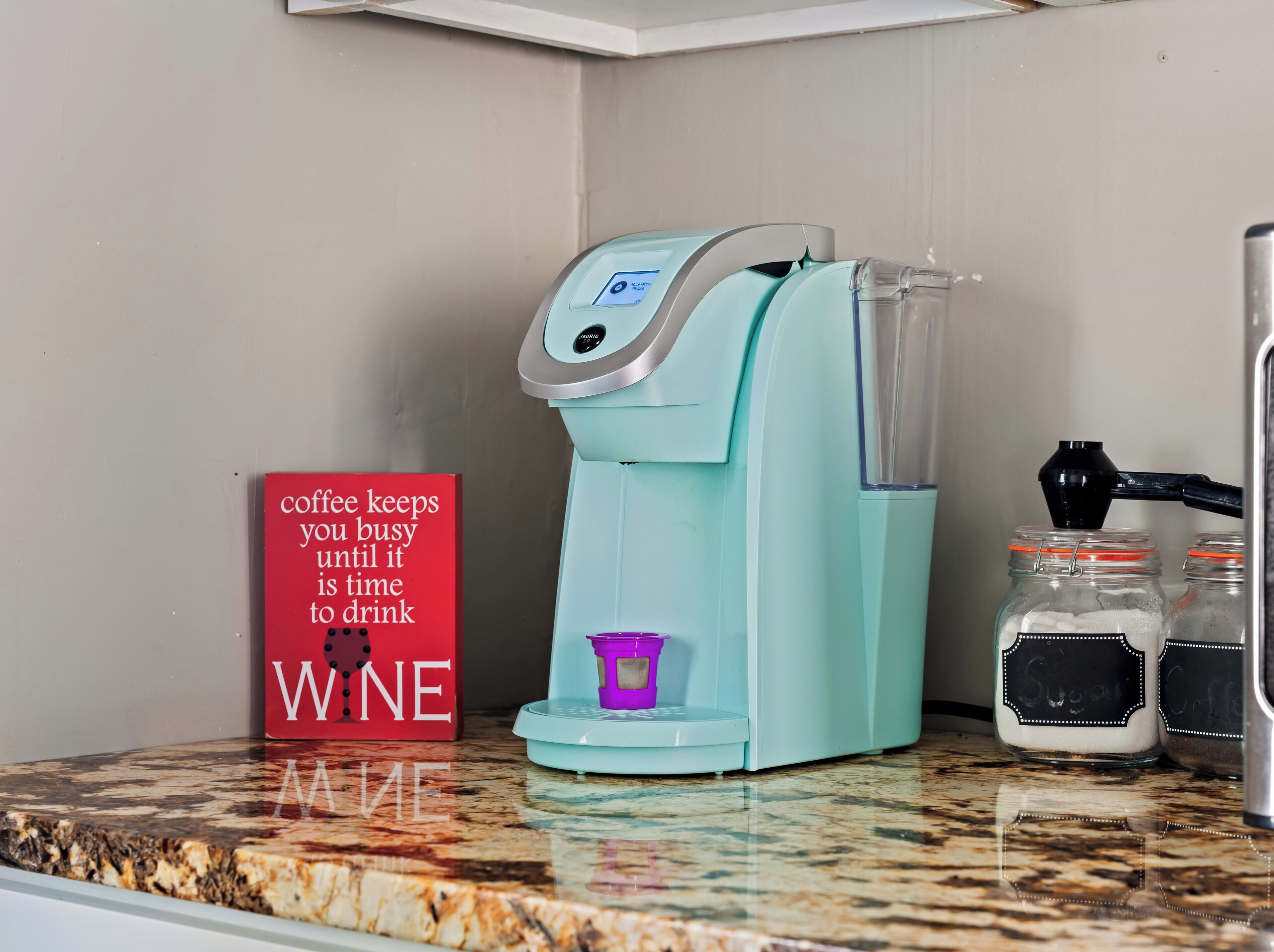 Eine Kaffeemaschine auf einer Küchenarbeitsplatte mit einem roten Schild, auf dem steht: "Kaffee hält dich beschäftigt, bis es Zeit für Wein ist."