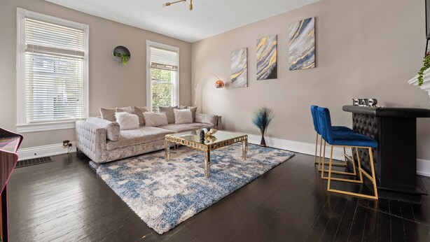 Ein Wohnzimmer mit einem beige farbenen Sofa, einem Glastisch, einem blau-weißen Teppich, einer schwarzen Bar mit goldenen Akzenten und einer modernen Chandelier.