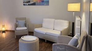 Ein gemütliches Wohnzimmer mit einer Couch, zwei Sesseln, einem runden Hocker, einer Stehlampe und einem Bild an der Wand.