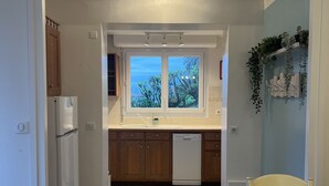 Eine Küche mit Holzausstattung, einem weißen Kühlschrank, einem Geschirrspüler und einem Fenster mit Blick auf Bäume. Ein Esstisch mit Stühlen und einer Tischdecke mit Streifenmuster.