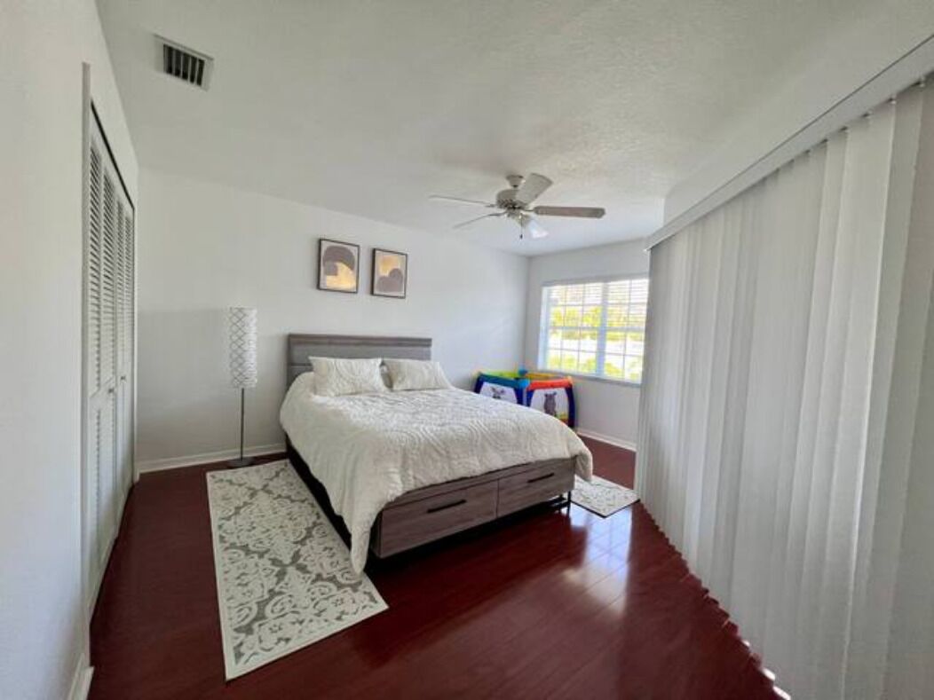 Quarto com cama, ventoinha no teto, janela com persianas e mesa de cabeceira com abajur.