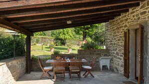 An outdoor patio with a stone table and wooden chairs, a stone wall, and a view of a garden. -  Il Mulino Di Vogognano by Geco Vacation Rentals (Subbiano)