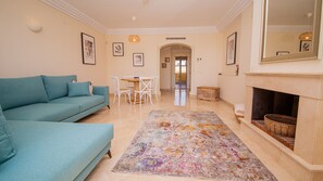 A living room with a sofa, a dining table with chairs, a fireplace, and a hallway leading to another room.