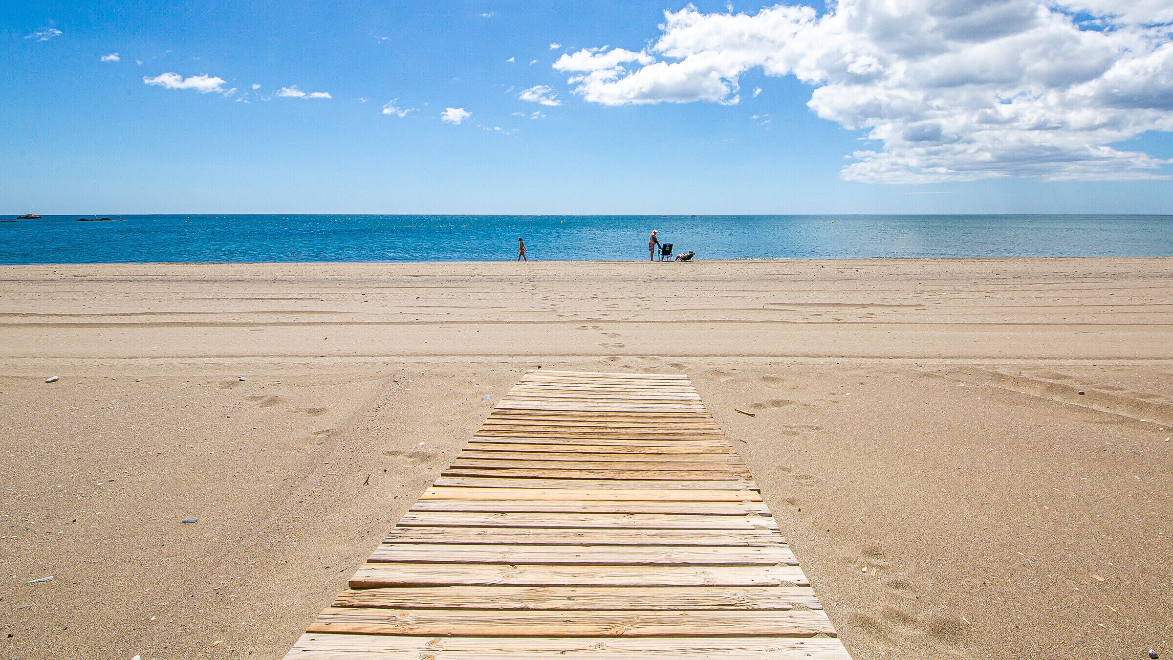 Een houten vlonder die naar een strand leidt, met helderblauw water en enkele mensen in de verte.