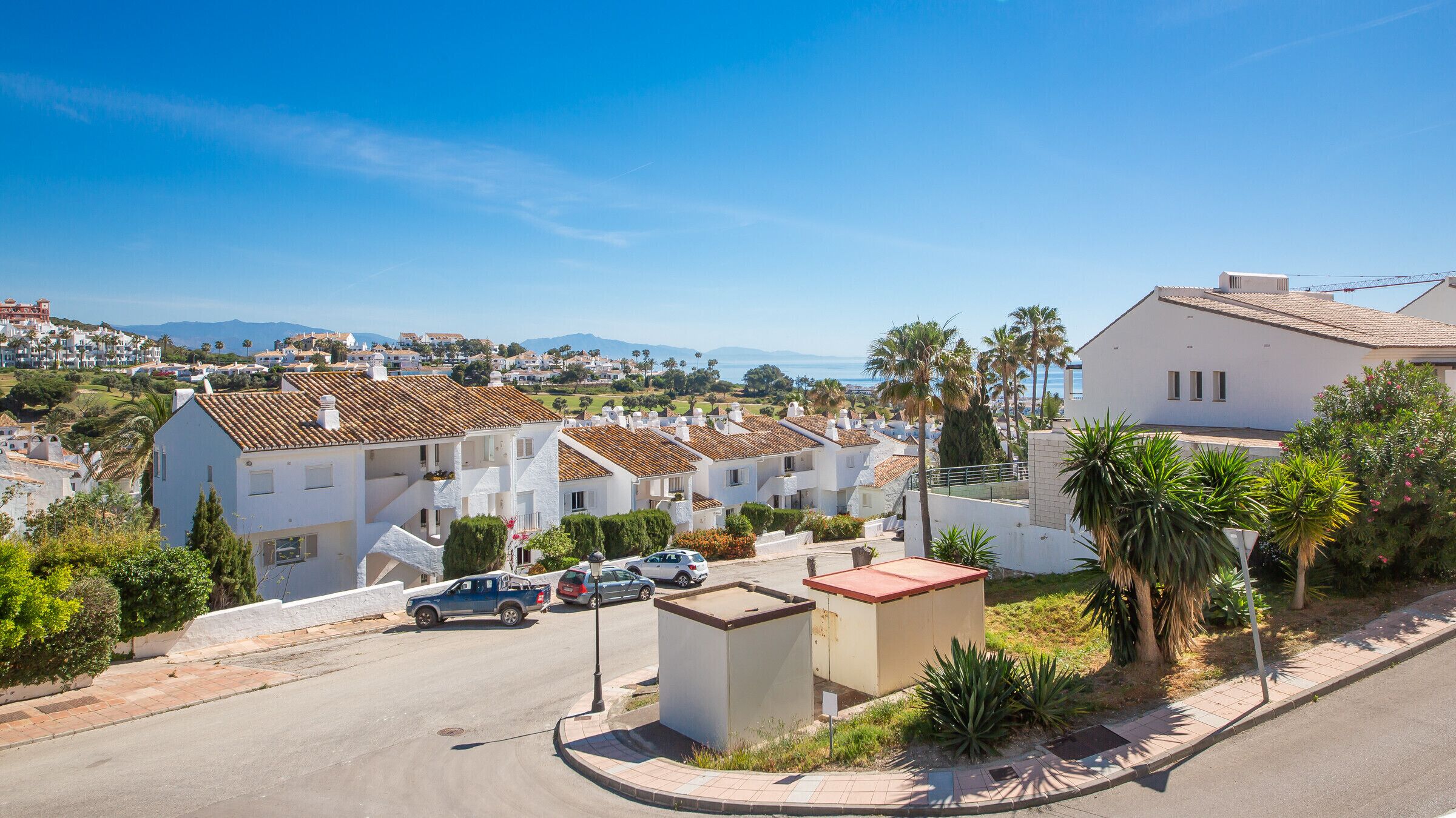 A residential street with white houses, a small utility box, and parked cars.