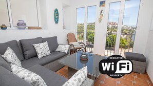 A furnished living room with a grey sofa, patterned cushions, a wicker coffee table, and a view of a cityscape through large sliding doors. - Terrazas de los Hidalgos F21A (Manilva)