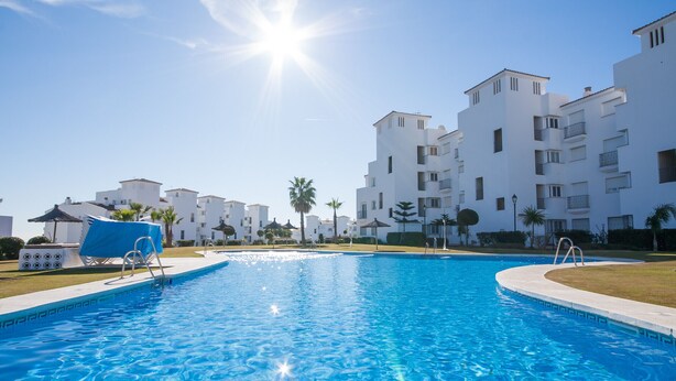 A swimming pool with clear blue water, surrounded by white buildings with balconies and palm trees. - Terrazas de los Hidalgos F21A (Manilva)