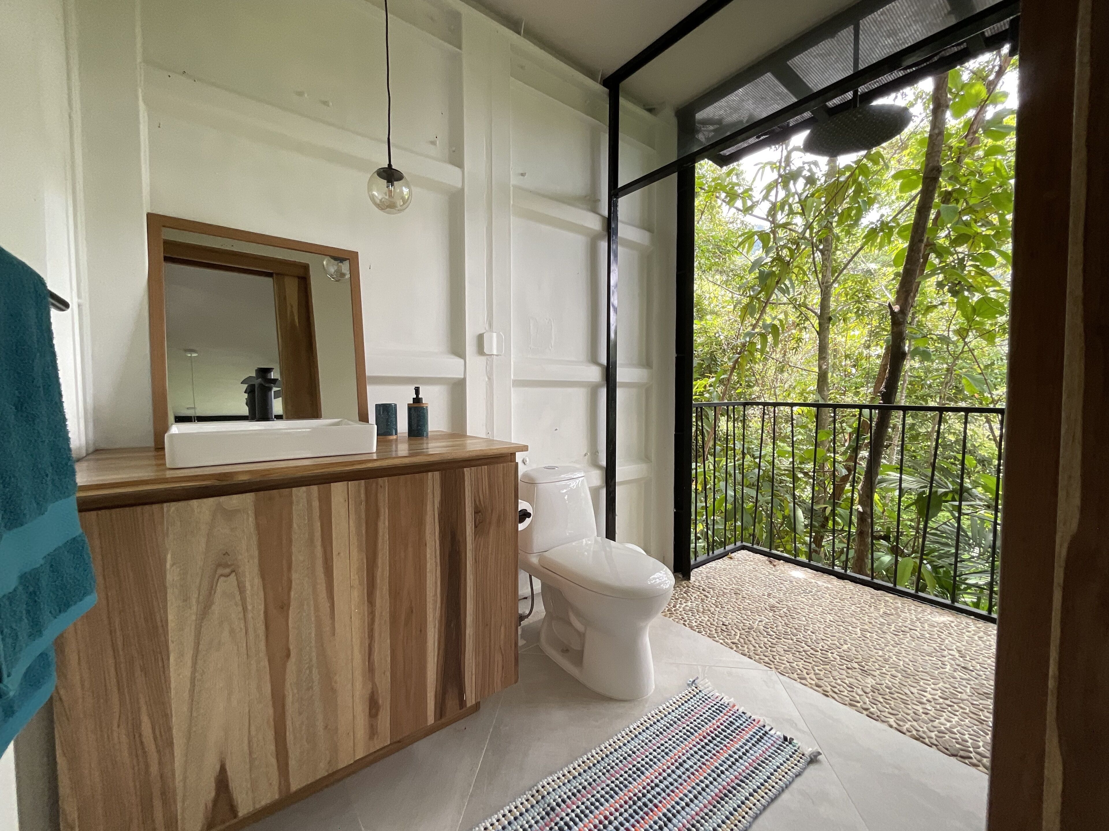 Ein Badezimmer mit Waschtisch aus Holz, Toilette und großem Fenster mit Balkon und Blick auf Bäume.