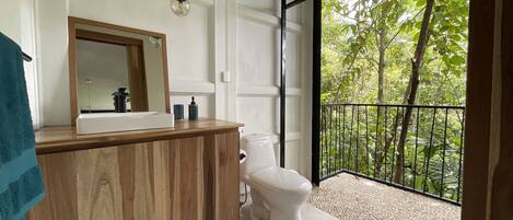 Ein Badezimmer mit Waschtisch aus Holz, Toilette und großem Fenster mit Balkon und Blick auf Bäume.