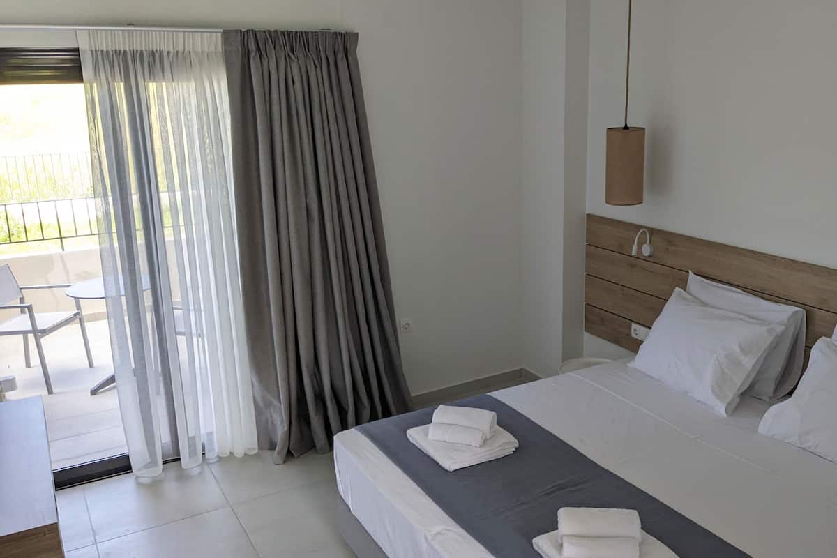 A hotel room with a bed, a bedside table, a chair, and a view of the outdoors through a window with curtains.
