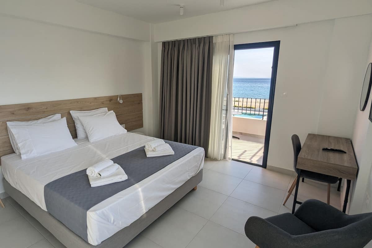 A hotel room with a bed, a desk, a chair, and a view of the sea through a large window.