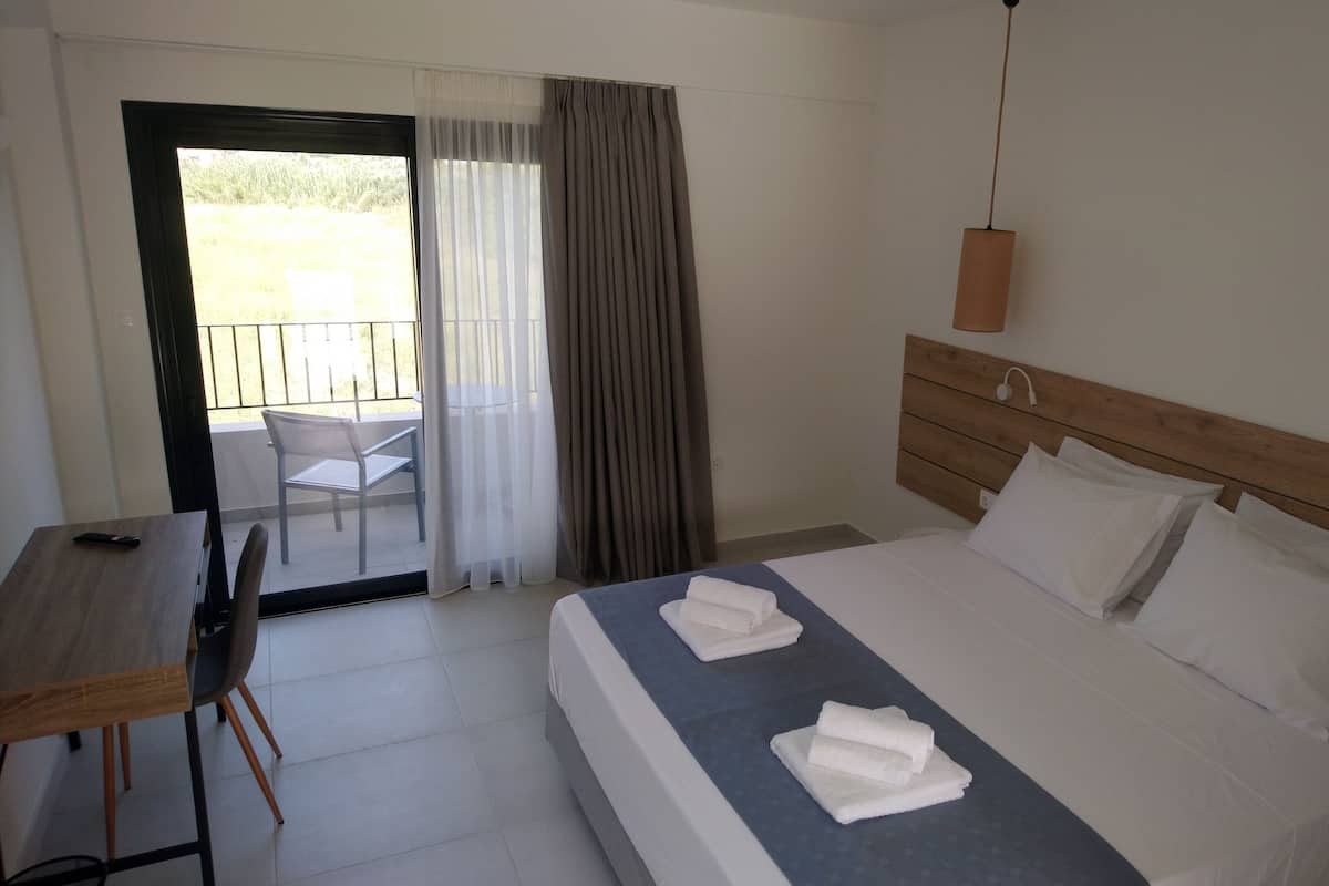 A hotel room with a bed, a desk, and a chair. There is a balcony with a chair outside the room.