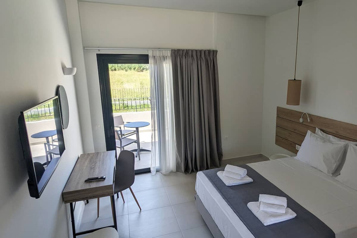 A hotel room with a bed, a desk with a chair, a TV, and a balcony with a table and chairs.