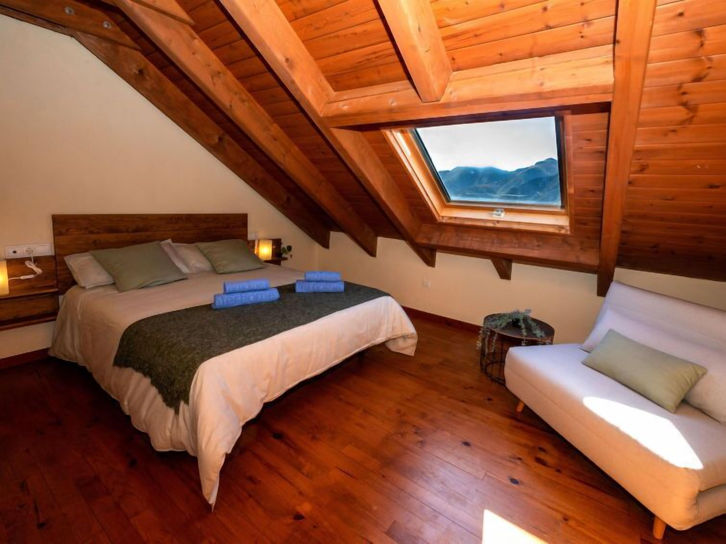 Une chambre confortable située sous les toits, avec un plafond en bois, une grande lucarne, un lit où les serviettes sont soigneusement pliées, une table de chevet avec une lampe et une petite table ronde agrémentée d’une plante.
