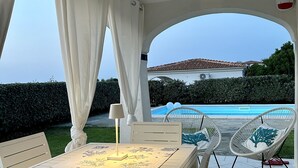 A covered patio with a table and chairs, a lamp, and a view of a swimming pool and building in the background.