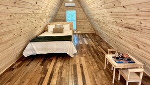 Une chambre mansardée chaleureuse avec des murs et un plancher en bois, un lit recouvert d’une courtepointe blanche, une petite table avec des livres et une fenêtre avec des stores.