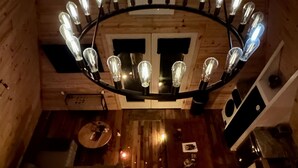 Un intérieur en bois avec un escalier menant à un deuxième étage, un lustre à plusieurs ampoules et un petit coin salon avec des coussins.
