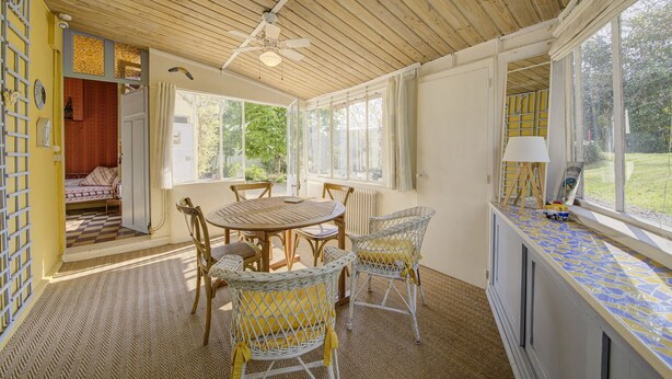 Une pièce baignée de lumière naturelle, avec un plafond en bois, une table à manger entourée de chaises, une fenêtre avec des rideaux et une petite table sur laquelle se trouve une lampe.