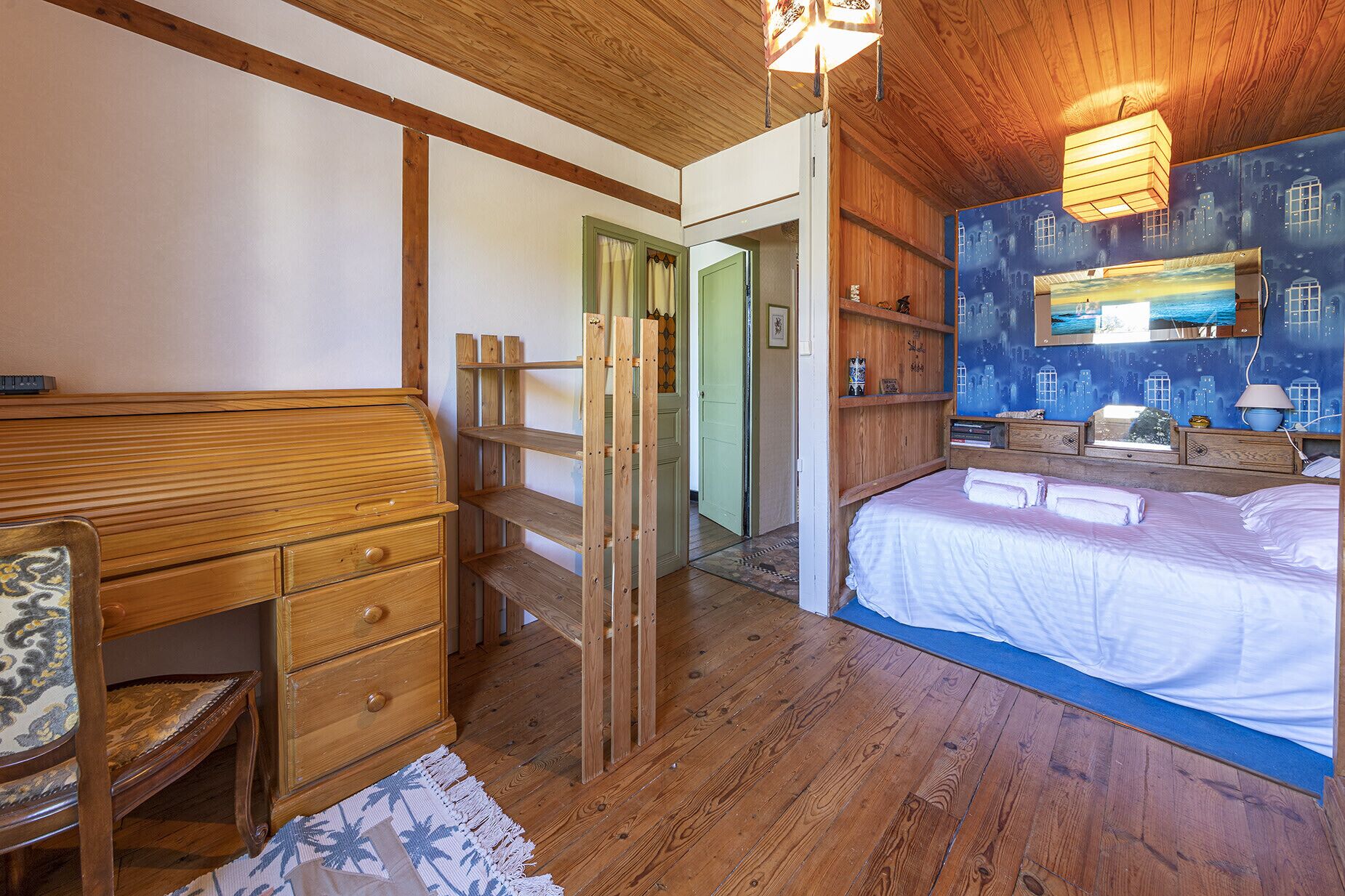Quarto com cama de madeira, mesa de cabeceira, cômoda e um mural com vista da cidade.
