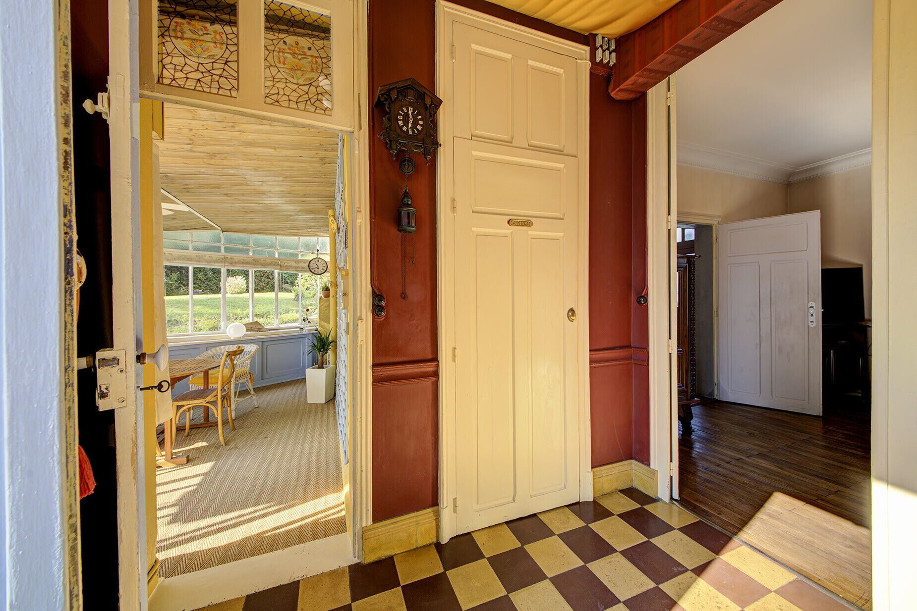 Un pasillo con piso a cuadros, una puerta blanca con placa y vista a una habitación con ventana y una silla de mimbre.