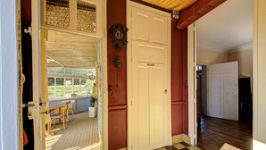 Un couloir doté d’un sol à carreaux, une porte blanche avec une plaque nominative, et une vue sur une pièce avec une fenêtre et un fauteuil en osier.