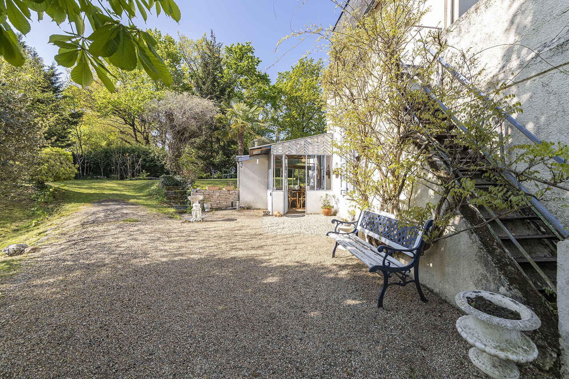 A gravel patio with a bench, a small shed, and a garden area with trees and shrubs.