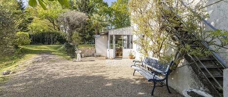 A gravel patio with a bench, a small shed, and a garden area with trees and shrubs.