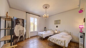Une chambre avec deux lits, un lustre, une grande fenêtre avec des rideaux, un plancher en bois et une bibliothèque ornée d’une statue.