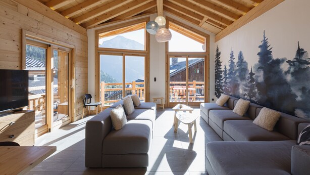 Ein großzügiger Wohnraum mit einer großen Couch, einem Couchtisch und einem Fernseher. Die Wände sind aus Holz und die hohe Decke hat sichtbare Balken. Große Fenster bieten einen Blick auf Berge und eine Holzterrasse außen.
