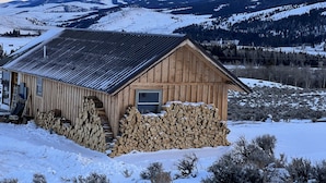 Een houten chalet met schuin dak, een raam en een deur, omringd door besneeuwde grond en bergen op de achtergrond.