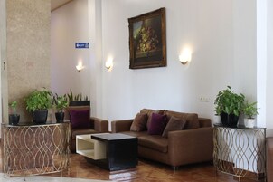 Un salón moderno con un sofá marrón, una mesa de centro y dos mesitas auxiliares con plantas en macetas. Hay un cuadro en la pared y una señal que indica la dirección de un ascensor.