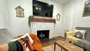 Ein Wohnzimmer mit Kamin aus weißem Mauerwerk, einem flachbildschirmfernseher darüber, einem grauen Sofa, einem Holztisch und einer grauen Couch mit Zierkissen.