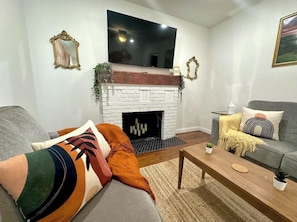 A living room with a white brick fireplace, a flat-screen TV mounted above it, a grey sofa, a wooden coffee table, and a grey couch with decorative pillows.