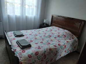 Una cama bien hecha con una colcha estampada con motivos florales y dos toallas dobladas, un cabecero de madera, una mesita de noche con una lámpara y una ventana con cortinas de gasa.