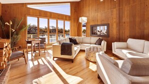 A spacious room with wooden walls, large windows, a sofa, a chair, a table with chess, and a lamp. - Waterfront | Minutes to US Open| Private Water Access (Hampton Bays)