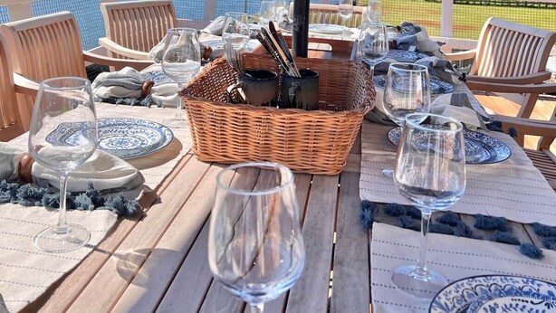 An outdoor dining area with a view of the water, a table set with plates and glasses, and a wicker basket. - Waterfront | Minutes to US Open| Private Water Access (Hampton Bays)
