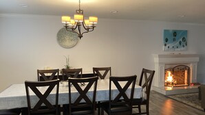 Ein Essbereich mit einem für sechs Personen gedeckten Tisch, umgeben von dunklen Holzstühlen. Eine Chandelüster hängt darüber, und ein Gemälde ist an der Wand zu sehen. Im Hintergrund ist ein Kamin erkennbar.