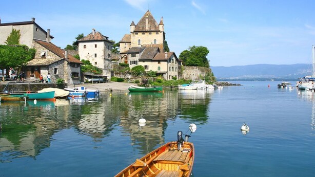 A calm lake with boats, a wooden rowboat in the foreground, and historic buildings on the shore. - Relais du Léman (Messery)