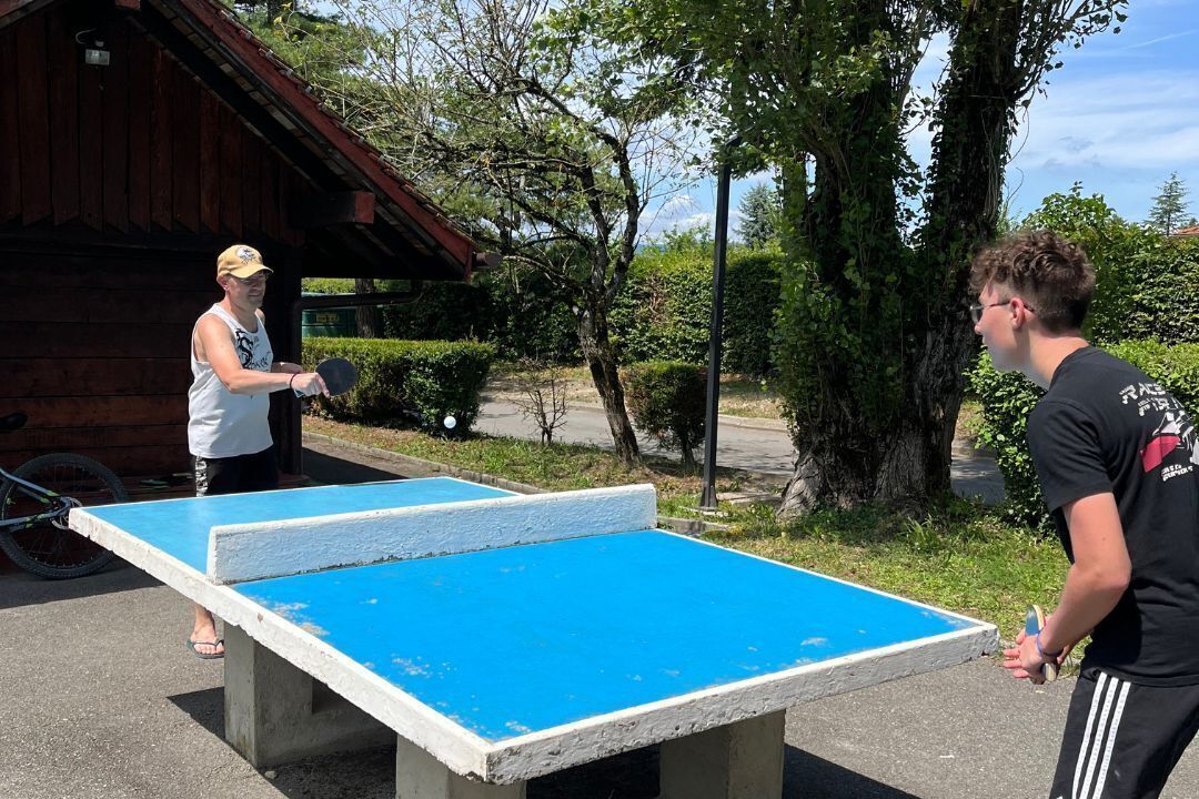 Dos personas jugando ping-pong al aire libre, con un edificio de madera y vegetación de fondo.