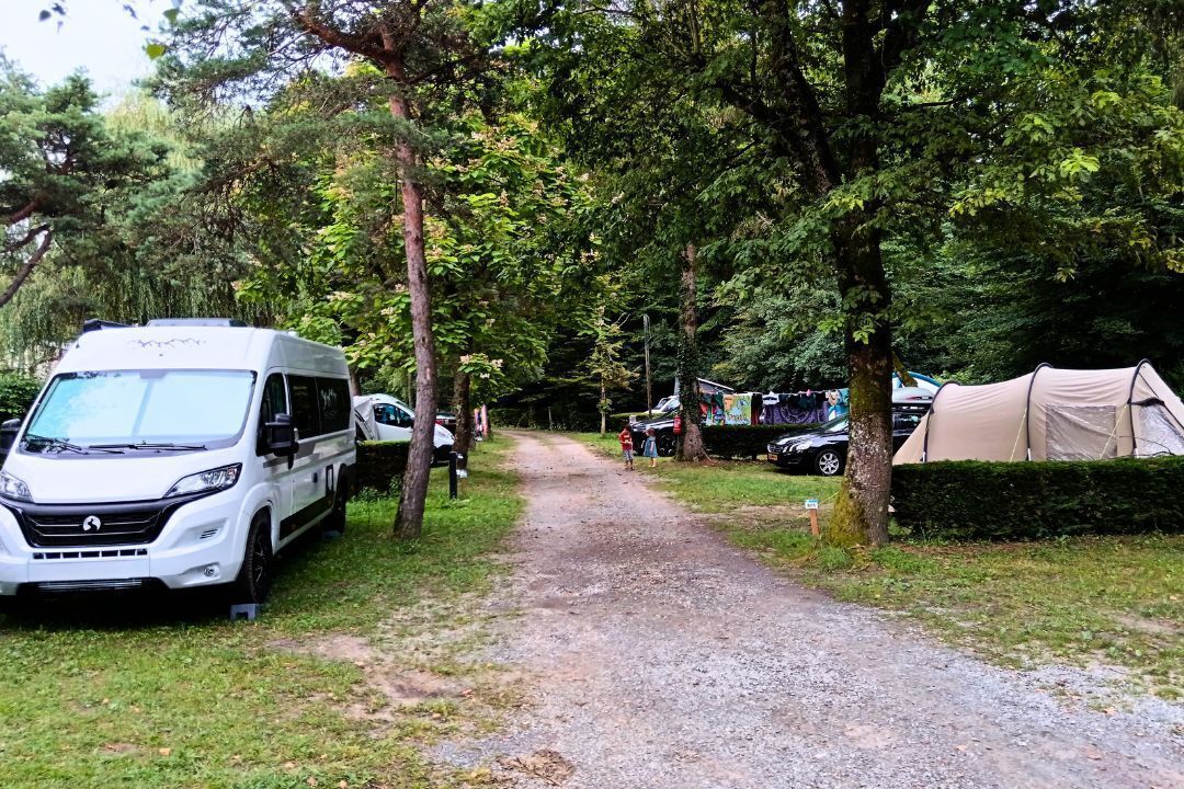 A gravel path flanked by trees, leading to a campsite with tents and a white RV parked on the side.