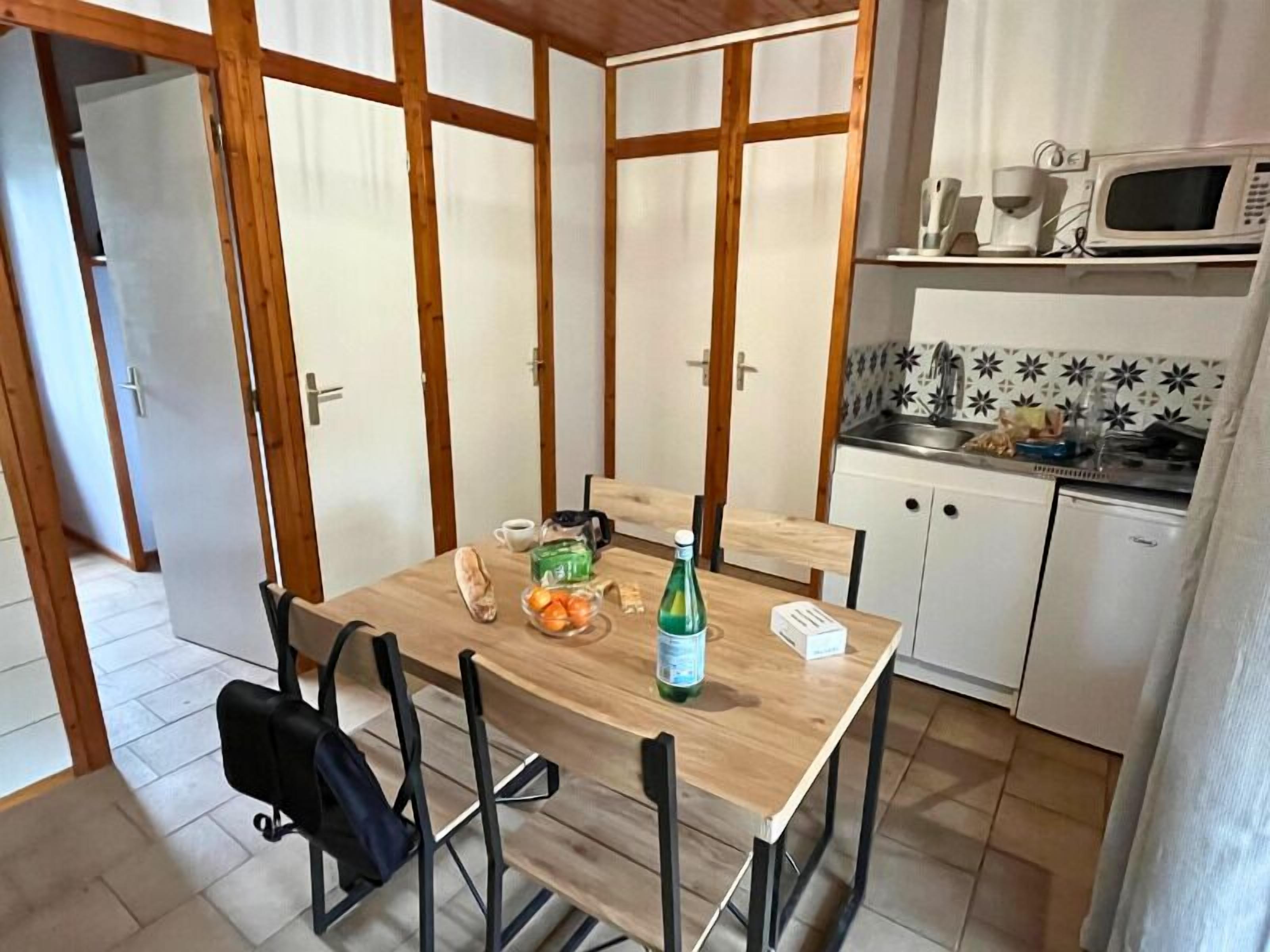 Een kleine keuken met een houten tafel, stoelen en basisvoorzieningen zoals een magnetron, spoelbak en kastjes.