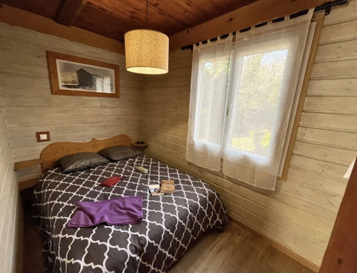 Habitació de cabana de fusta amb llit, llum penjant, finestra amb cortines lleugeres i un quadre al mur.