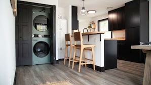 A compact kitchen with a washing machine, wooden chairs, and black cabinets.