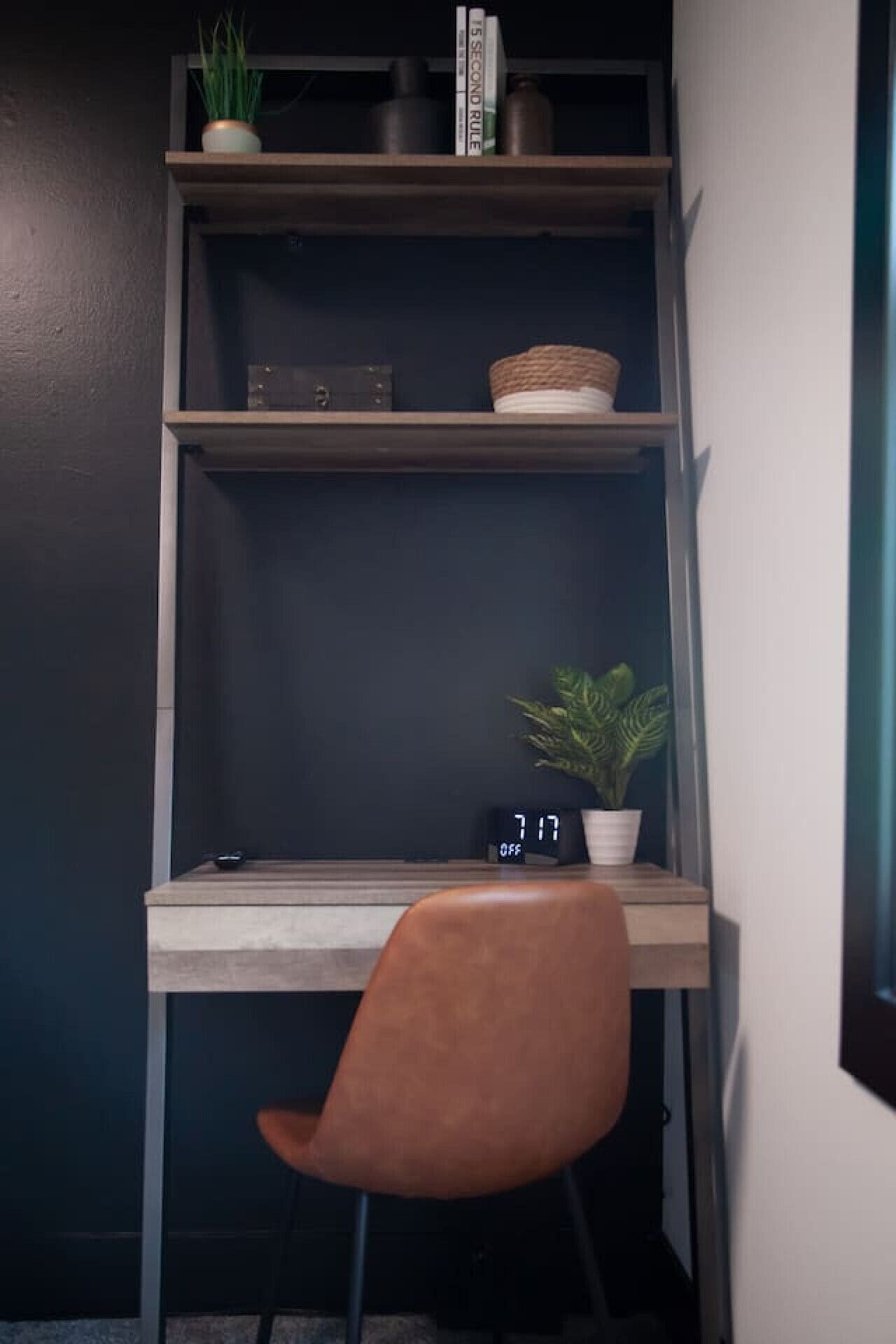 Un espace de travail d’angle avec un bureau en bois, une chaise marron, une petite plante en pot, une horloge numérique et une étagère contenant des livres et des objets décoratifs.