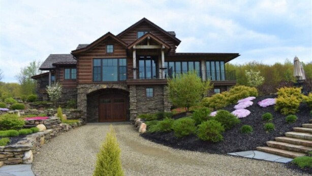 A large, two-story log cabin with a stone foundation, large windows, and a well-maintained garden with various shrubs and flowers.